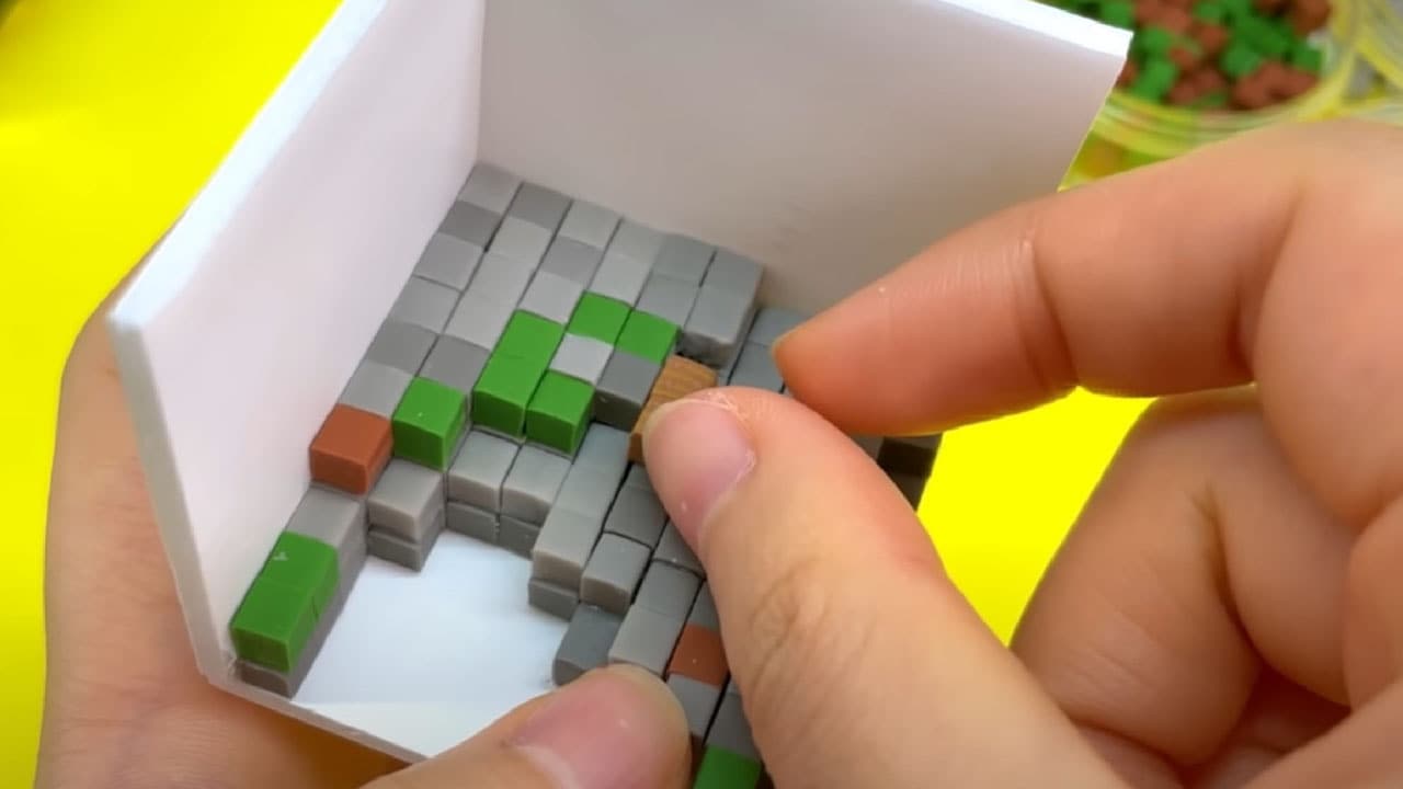 Photo of two hands against a yellow background holding white clay forming a corner of an open cube with a floor and two walls, and placing individual grey, green, and brown clay cubes resembling Minecraft blocks