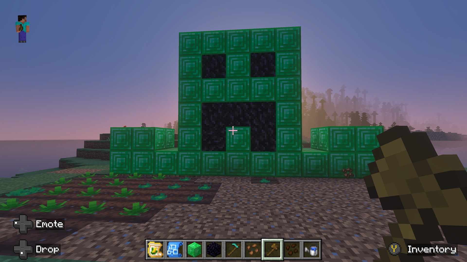 A large pixel-art face made from green and dark blocks stands beside farmland with newly planted crops at sunrise.