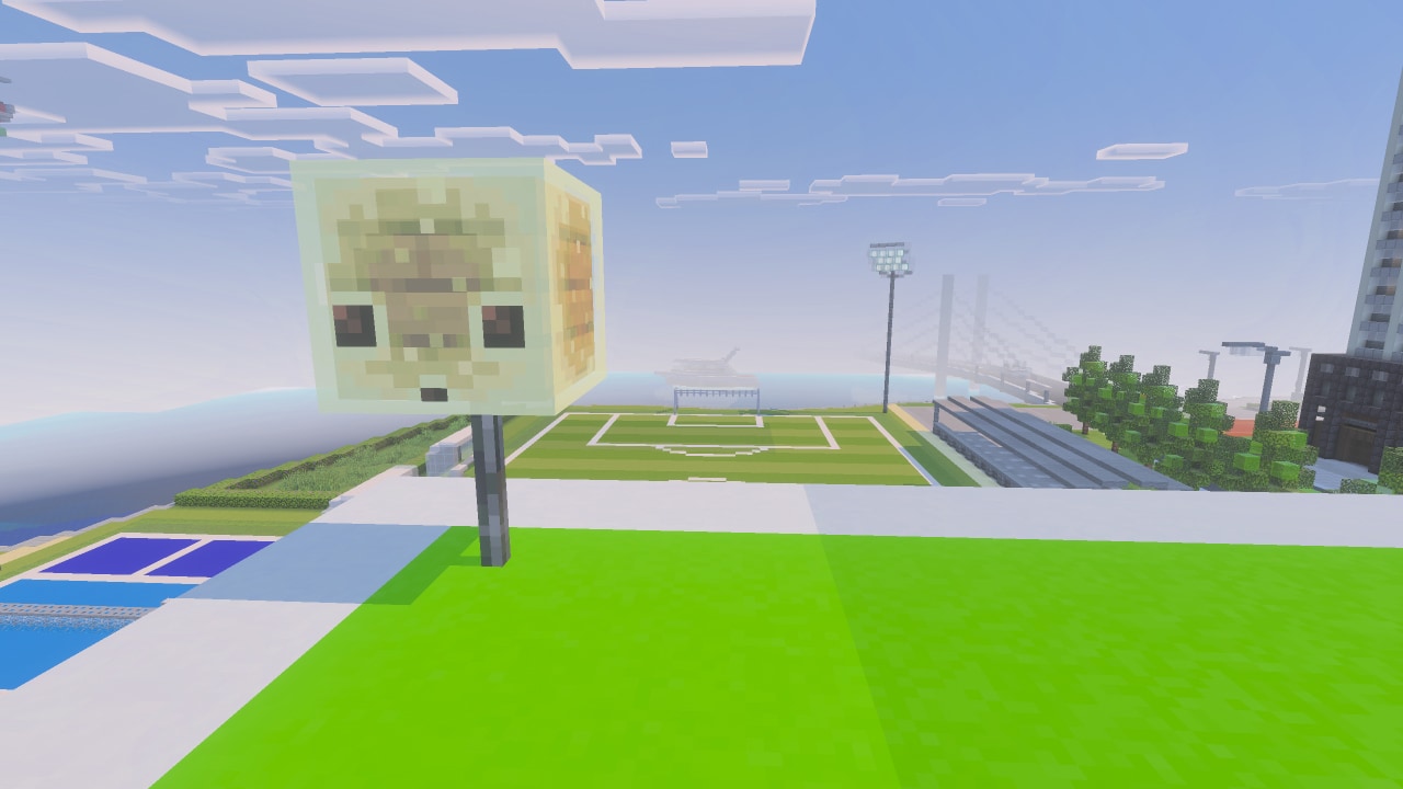 A giant sulfur cube on a pole overlooks a sports field beside a modern coastal cityscape.